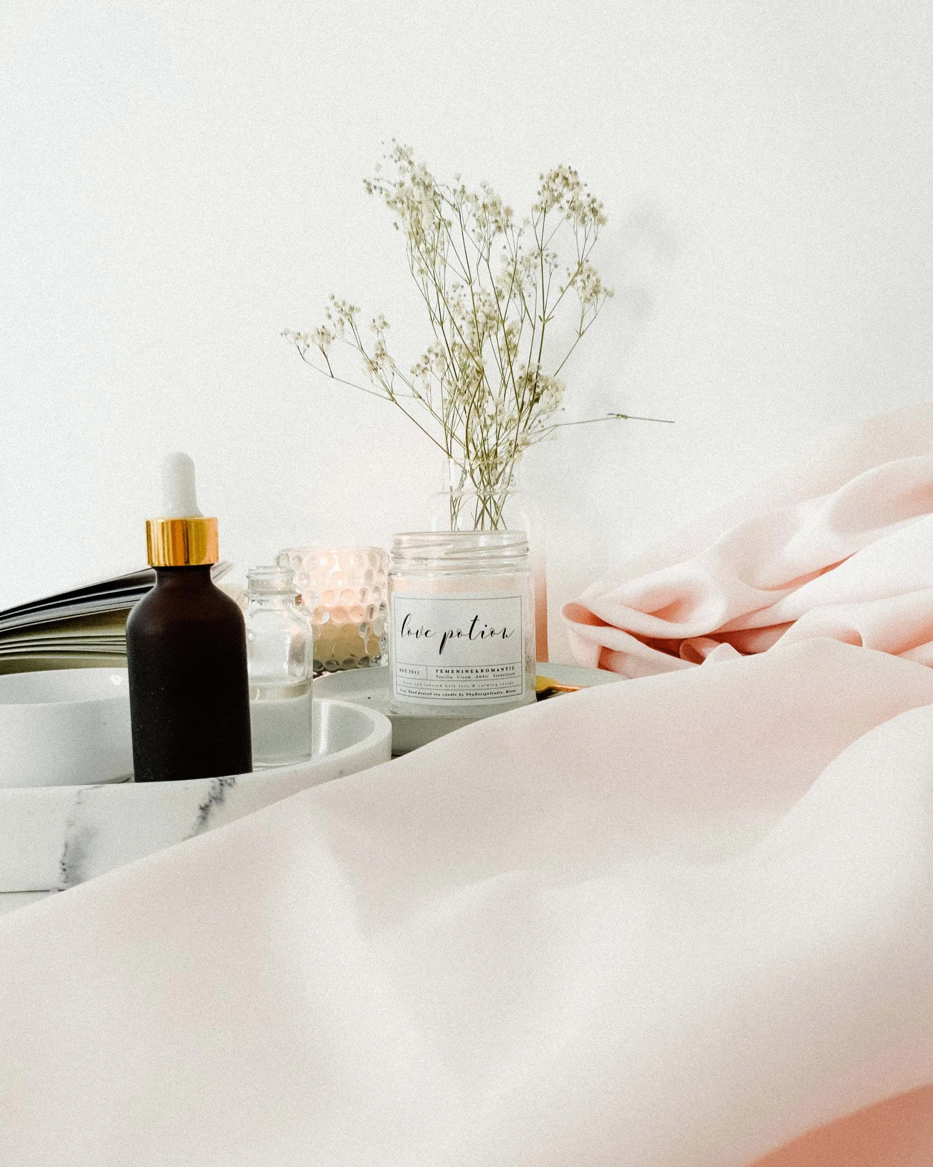 Studio flat-lay — amber dropper bottle, a candle, baby's-breath, pink silk
